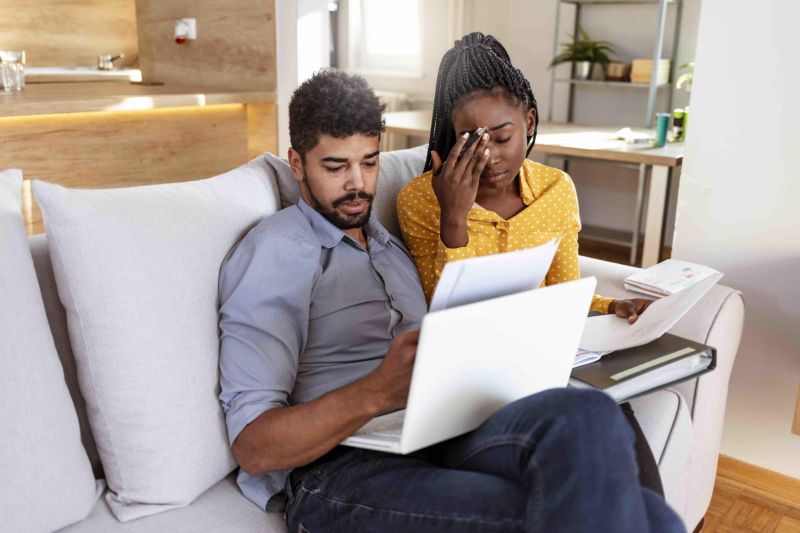 A man and a woman sitting on couch looking distressed with bills on hand