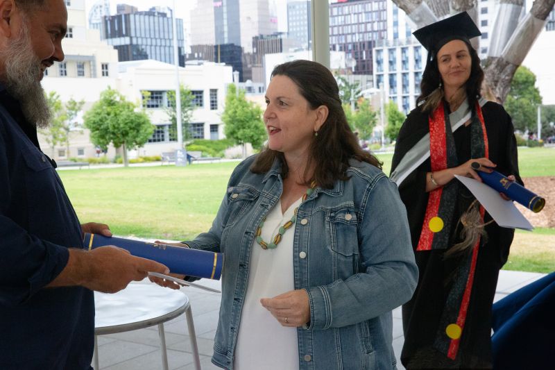 Man hands woman graduate diploma with another woman in the background